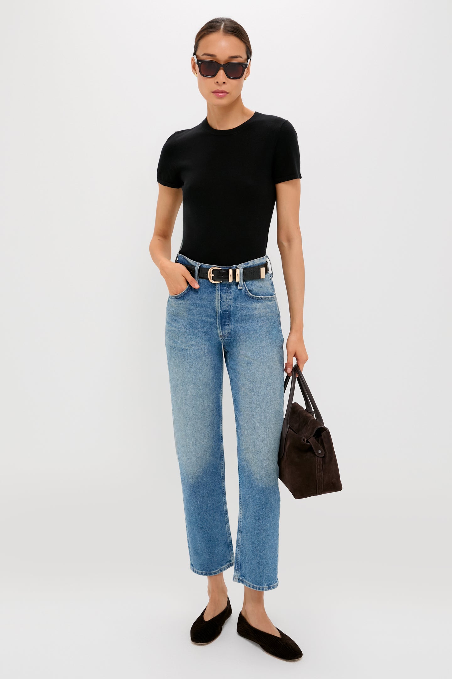 A woman in sunglasses, blue jeans, and black flats stands with a brown handbag against a white background, wearing a Tuckernuck Black Cashmere Silk Crewneck Short Sleeve Tee.