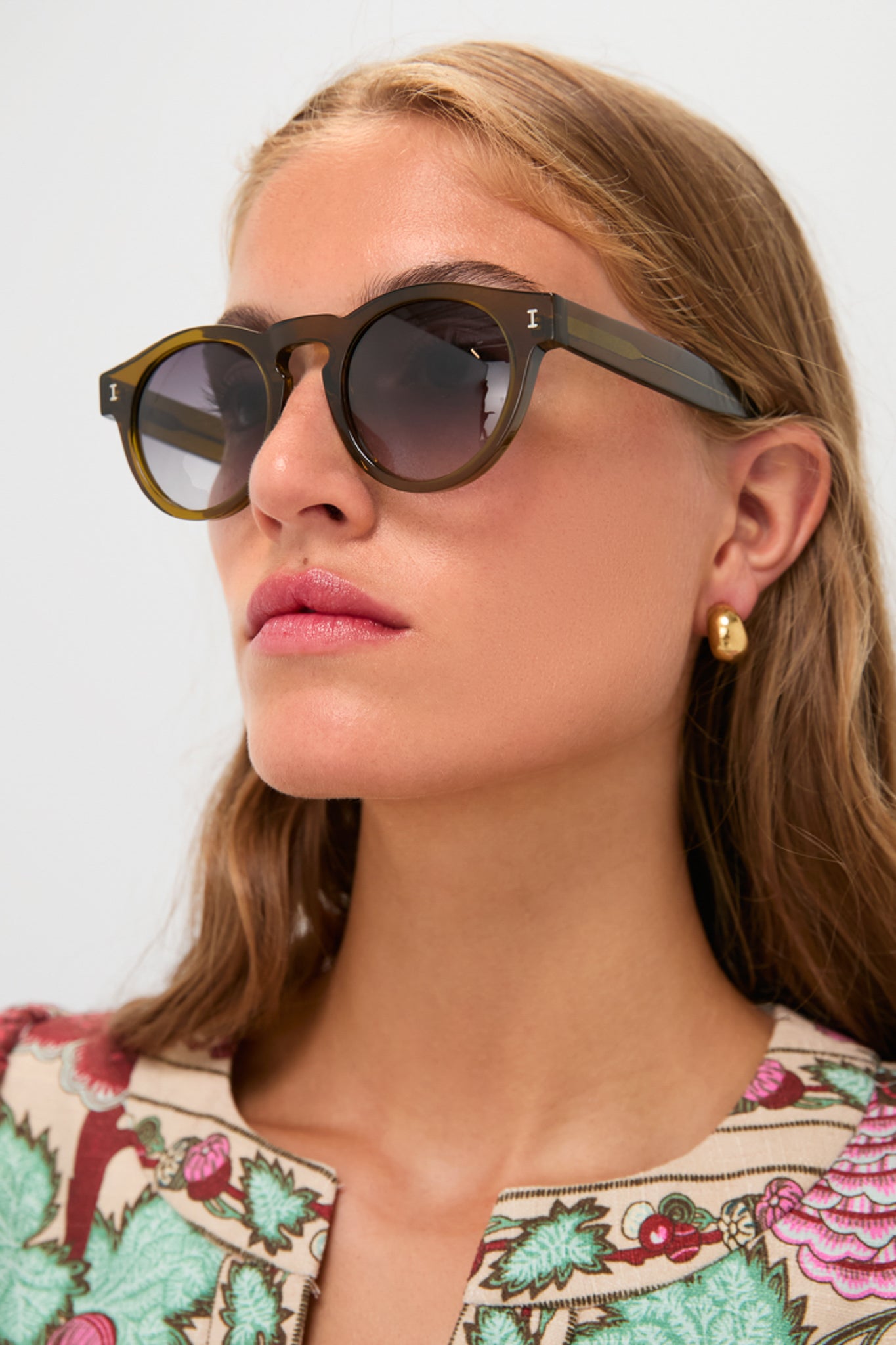 A woman wearing Illesteva round sunglasses, gold earrings, and a floral-patterned top is shown from the shoulders up against a plain background. The stylish sunglasses are handmade in Italy for timeless elegance.