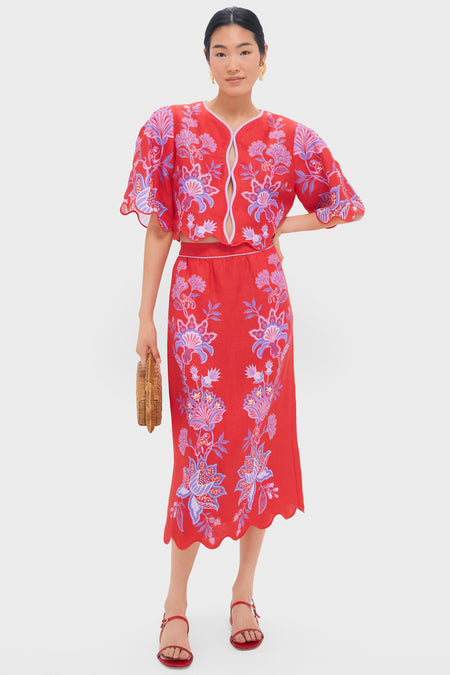 A woman wears a red floral Farm Rio two-piece with short puff sleeves, an A-line midi skirt with a scalloped hem. She accessorizes with a woven clutch and red sandals.
