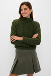 A woman models a Hyacinth House chunky green knit sweater and an olive green pleated skirt, standing with arms crossed and smiling.