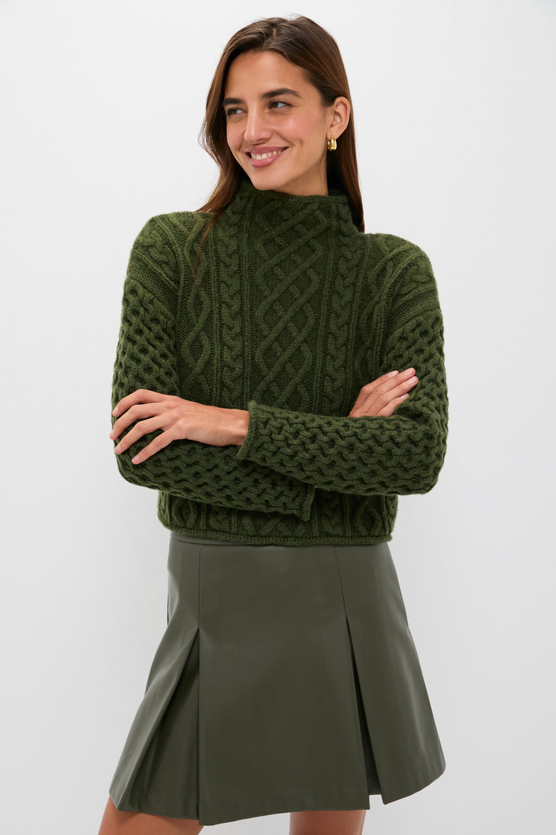 A woman models a Hyacinth House chunky green knit sweater and an olive green pleated skirt, standing with arms crossed and smiling.