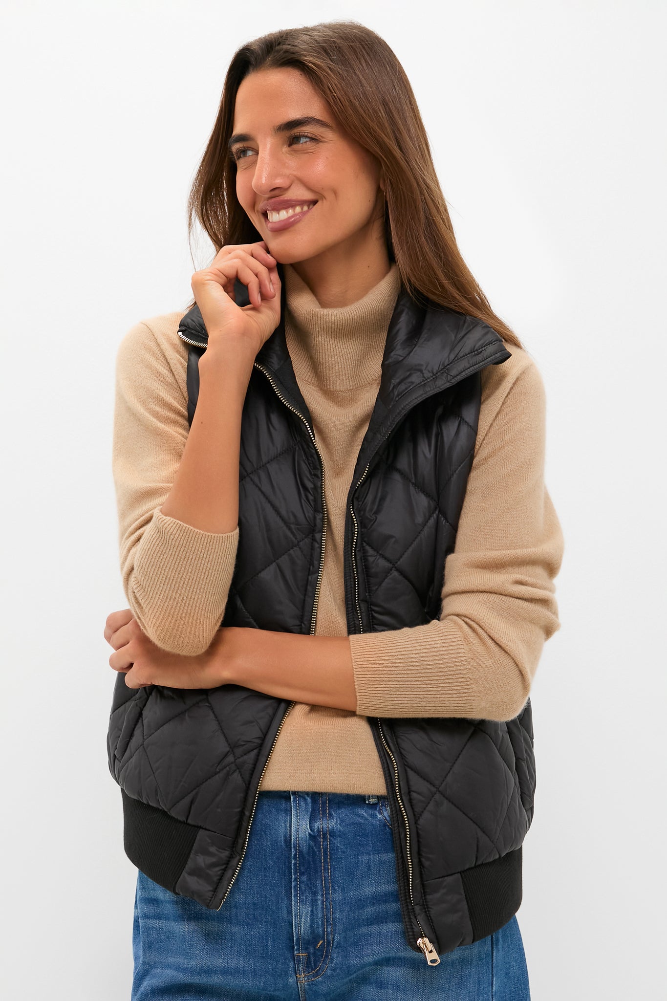 A woman stands with arms crossed, smiling in a Tuckernuck beige turtleneck sweater, blue jeans, and a black quilted vest—showcasing the perfect transitional outerwear look against a plain white background.