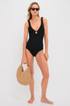 A woman wears a black crinkle one-piece swimsuit with a keyhole front, styled with a straw bag and sunglasses.