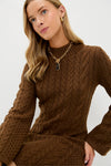 A person with long blonde hair wears a brown Hyacinth House Coffee Lucia cable knit dress, gold earrings, and a chunky gold chain necklace against a plain white background.
