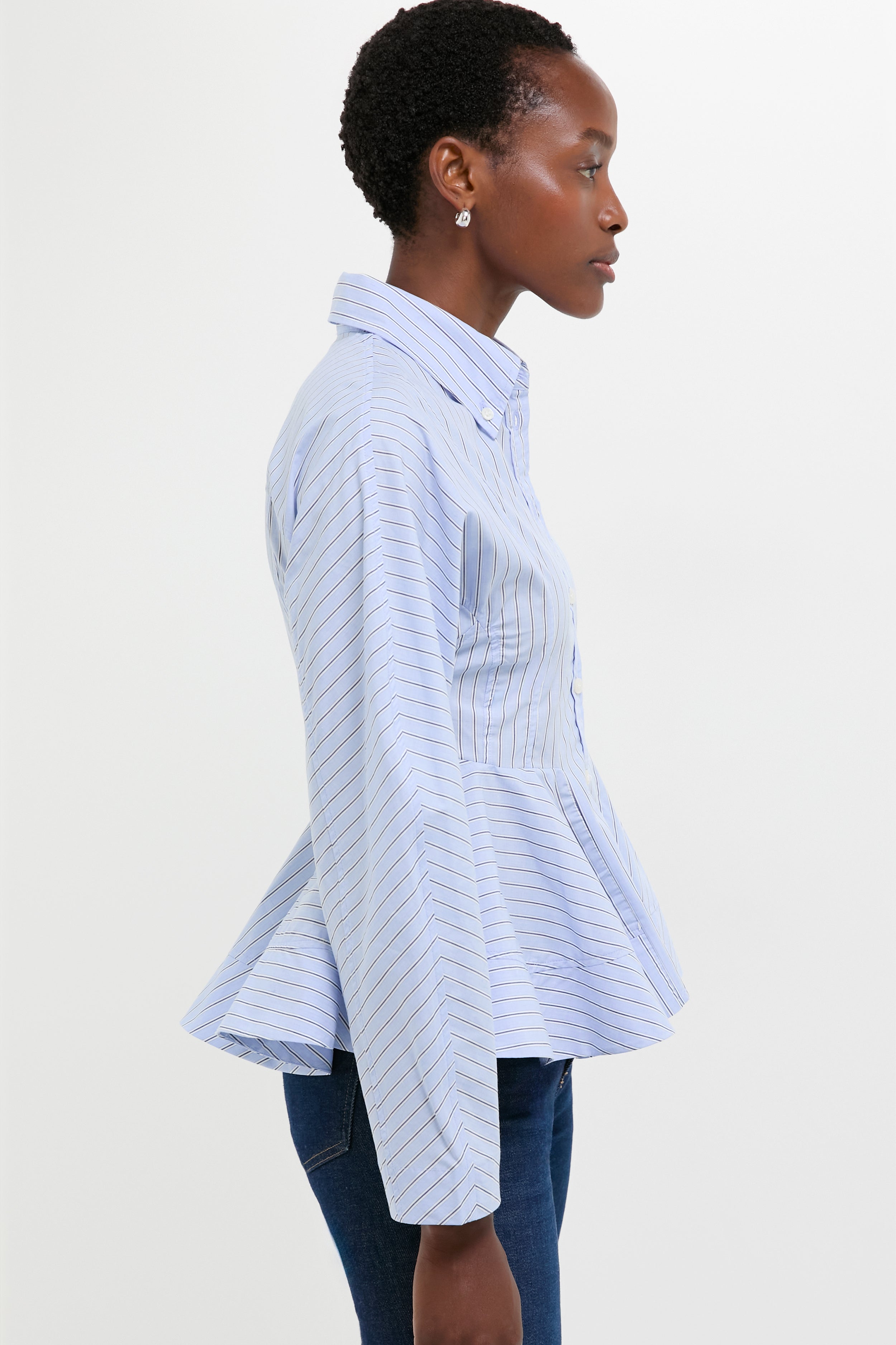 A woman wearing a GANNI blue and white striped poplin shirt with a peplum waist and dark jeans stands in profile against a plain white background.