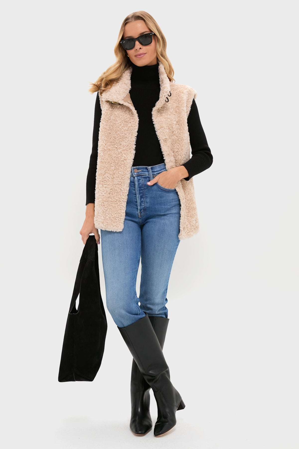 A woman stands against a plain background wearing a Tuckernuck beige sherpa vest, black high-neck turtleneck sweater, blue jeans, black knee-high boots, sunglasses, and holding a black bag.
