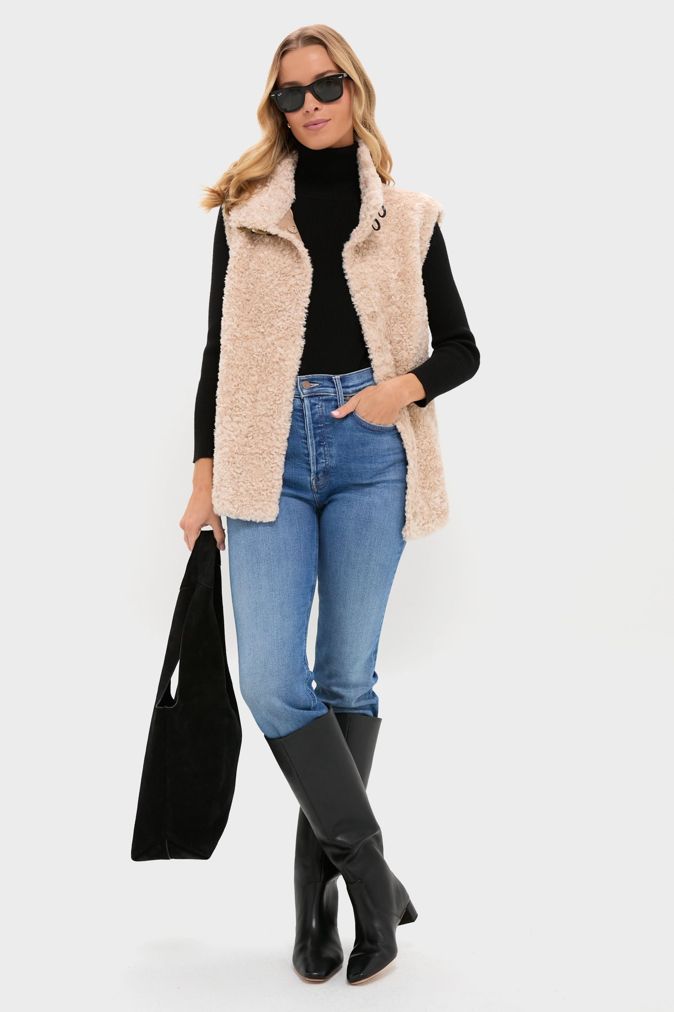 A woman stands against a plain background wearing a Tuckernuck beige sherpa vest, black high-neck turtleneck sweater, blue jeans, black knee-high boots, sunglasses, and holding a black bag.