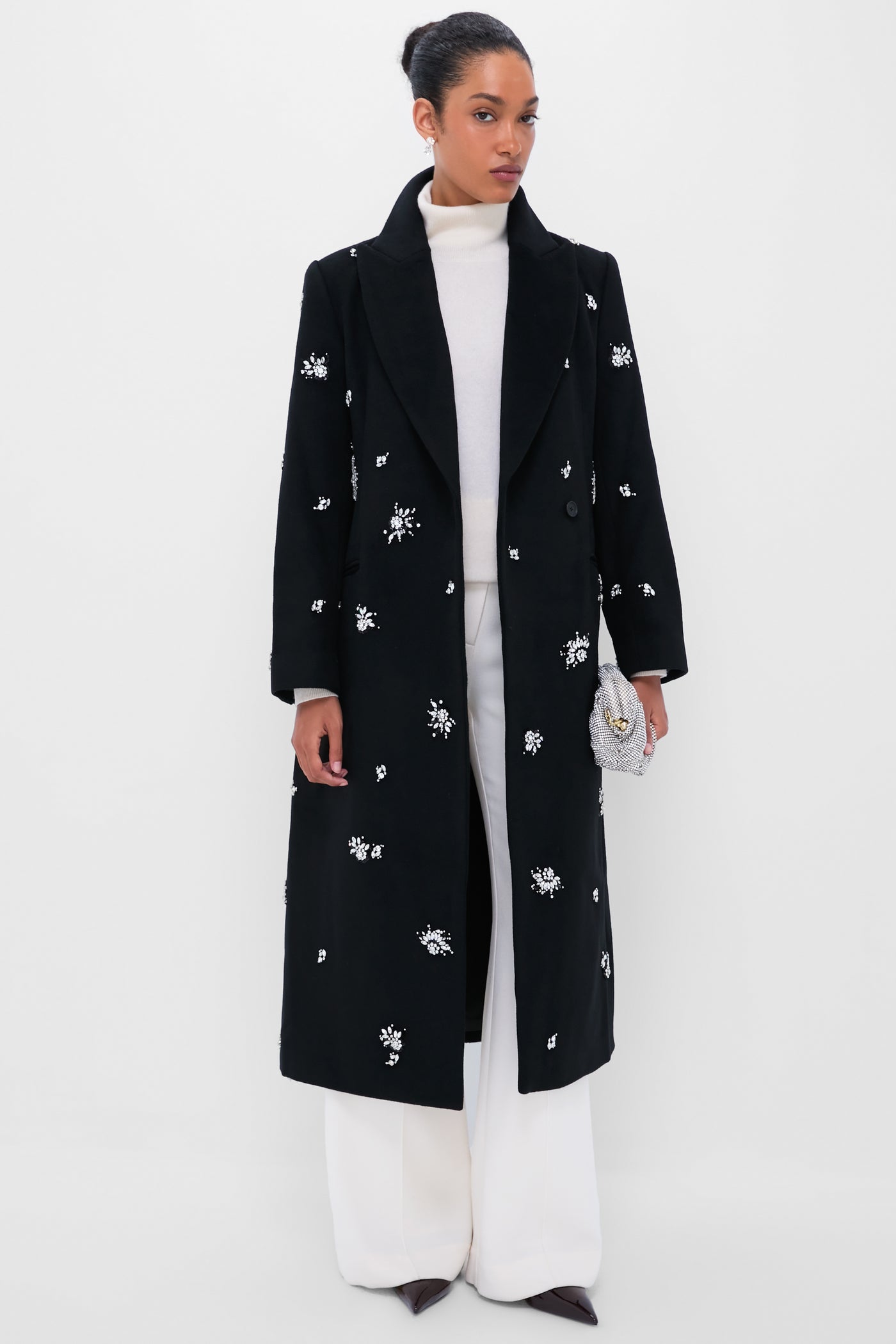 Model in a classic black overcoat with floral embellishments, wearing Crystal Pamina Earrings, embodying effortless elegance and fun, accessorized with a silvery clutch.