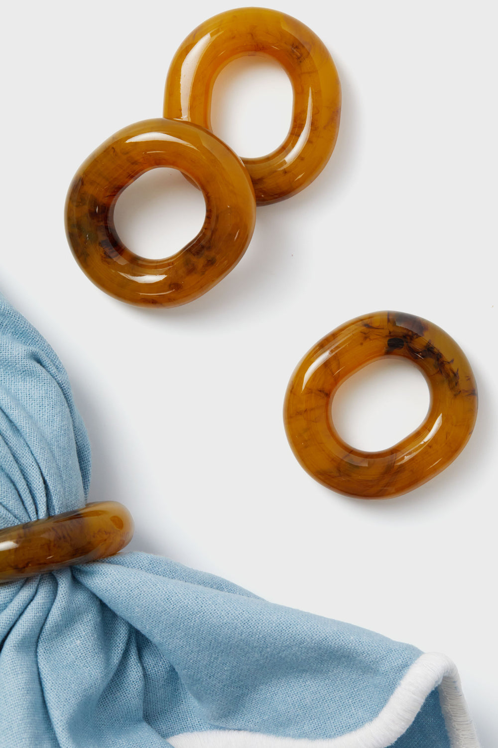 Four oval-shaped amber Tortoise Catena Link Napkin Rings by Tuckernuck Home, one displaying light blue fabric, are arranged on a white surface—ideal as hostess gifts or for entertaining.
