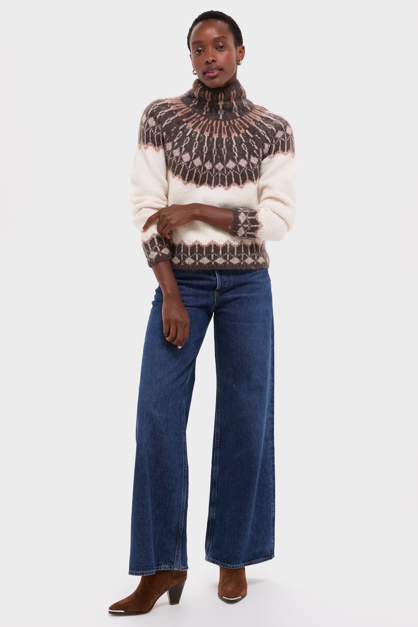 A person wears a Love the Label fair isle knit turtleneck sweater, wide-leg blue jeans, and brown heeled boots while standing against a plain white background.