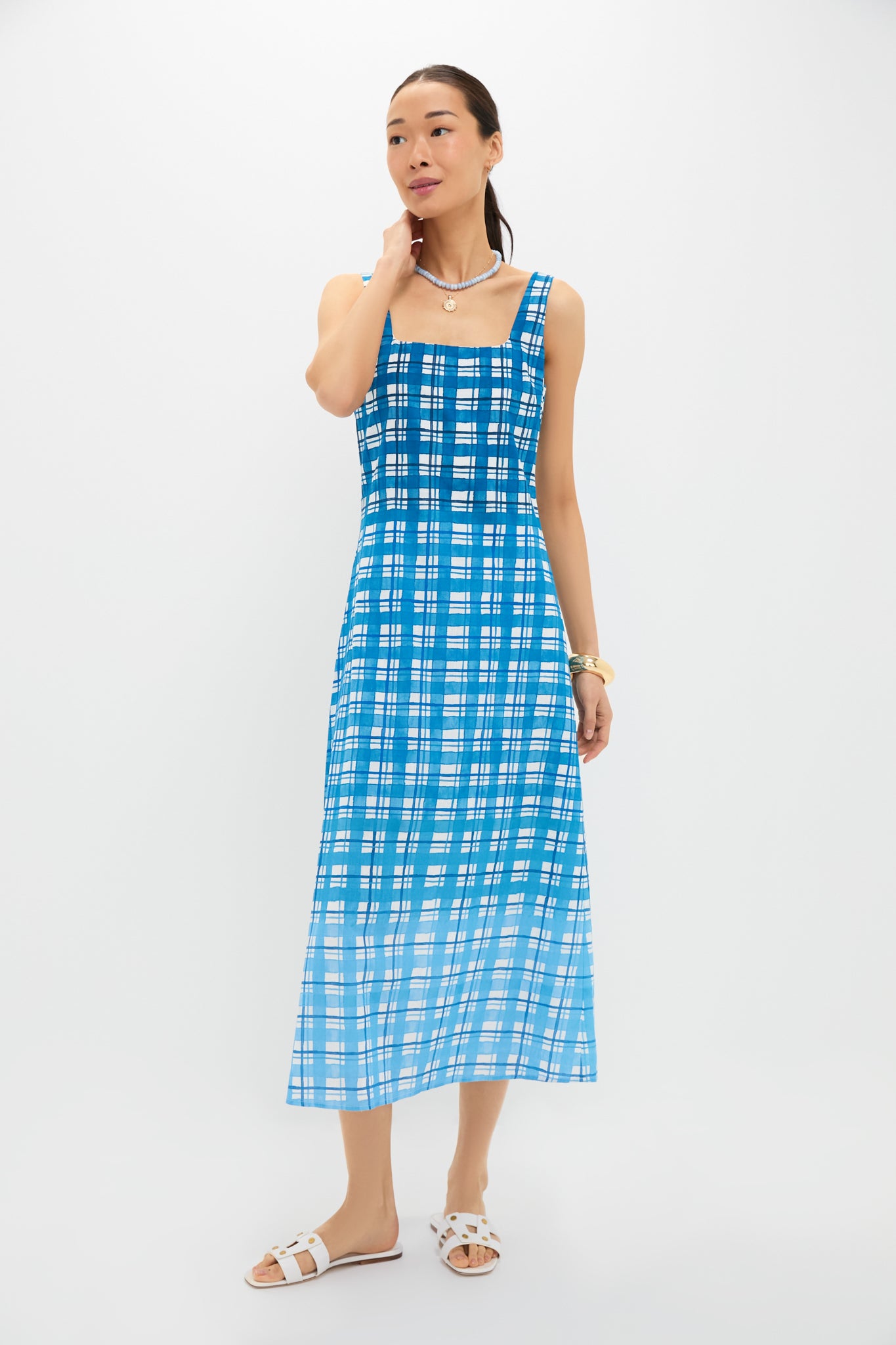 A woman in an Anna Cate blue and white plaid linen dress with a square neckline and white sandals stands in a minimalist setting.