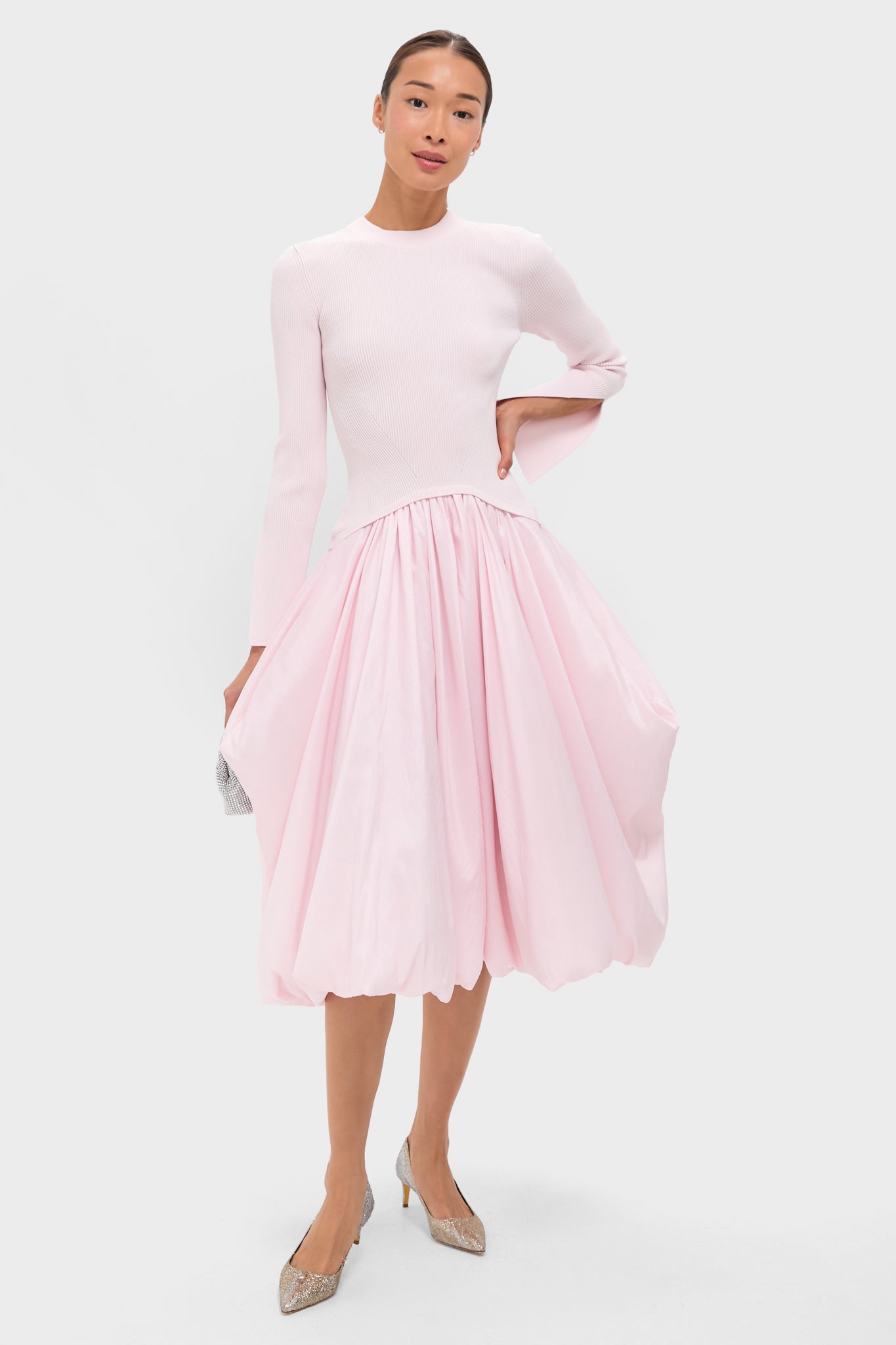 A woman in a SIMKHAI luxury ready-to-wear long-sleeved light pink dress with a fitted bodice and bubble-hem skirt, silver heels, and a small clutch stands against a plain background.