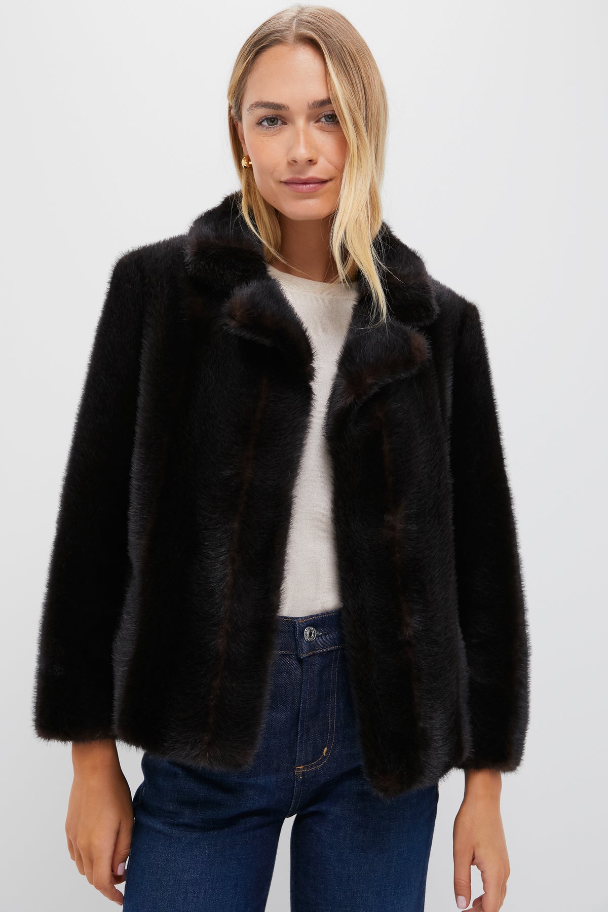 A woman models a Tuckernuck dark faux fur coat layered over a light top and blue jeans, posing against a plain white background.