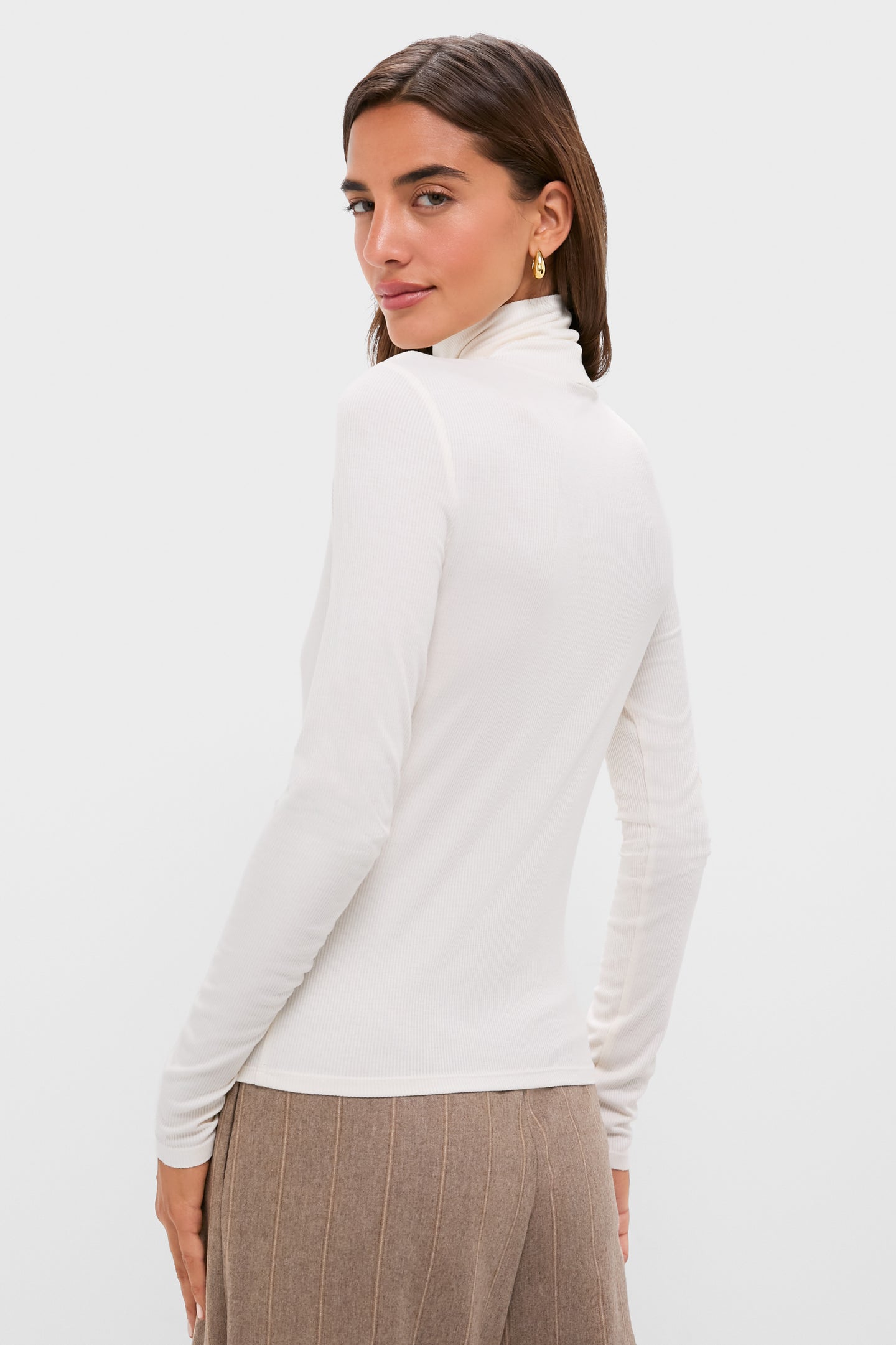 A woman with brown hair wears a Tuckernuck white ribbed turtleneck long-sleeve top and beige pants, standing with her back to the camera and turning her head slightly.