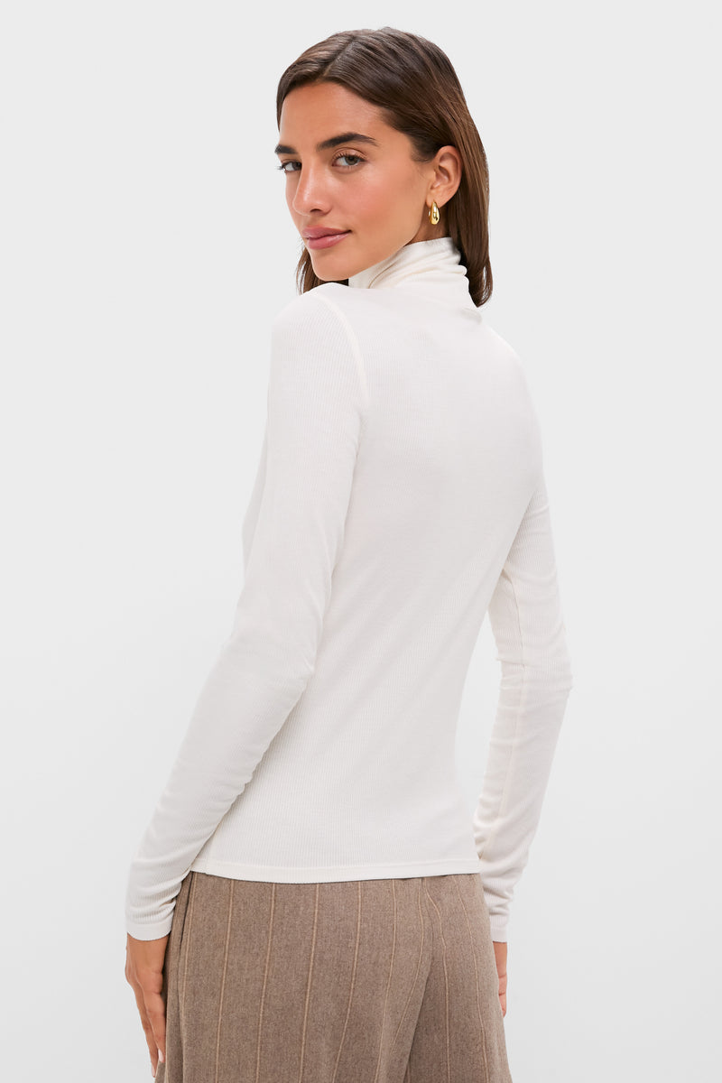 A woman with brown hair wears a Tuckernuck white ribbed turtleneck long-sleeve top and beige pants, standing with her back to the camera and turning her head slightly.