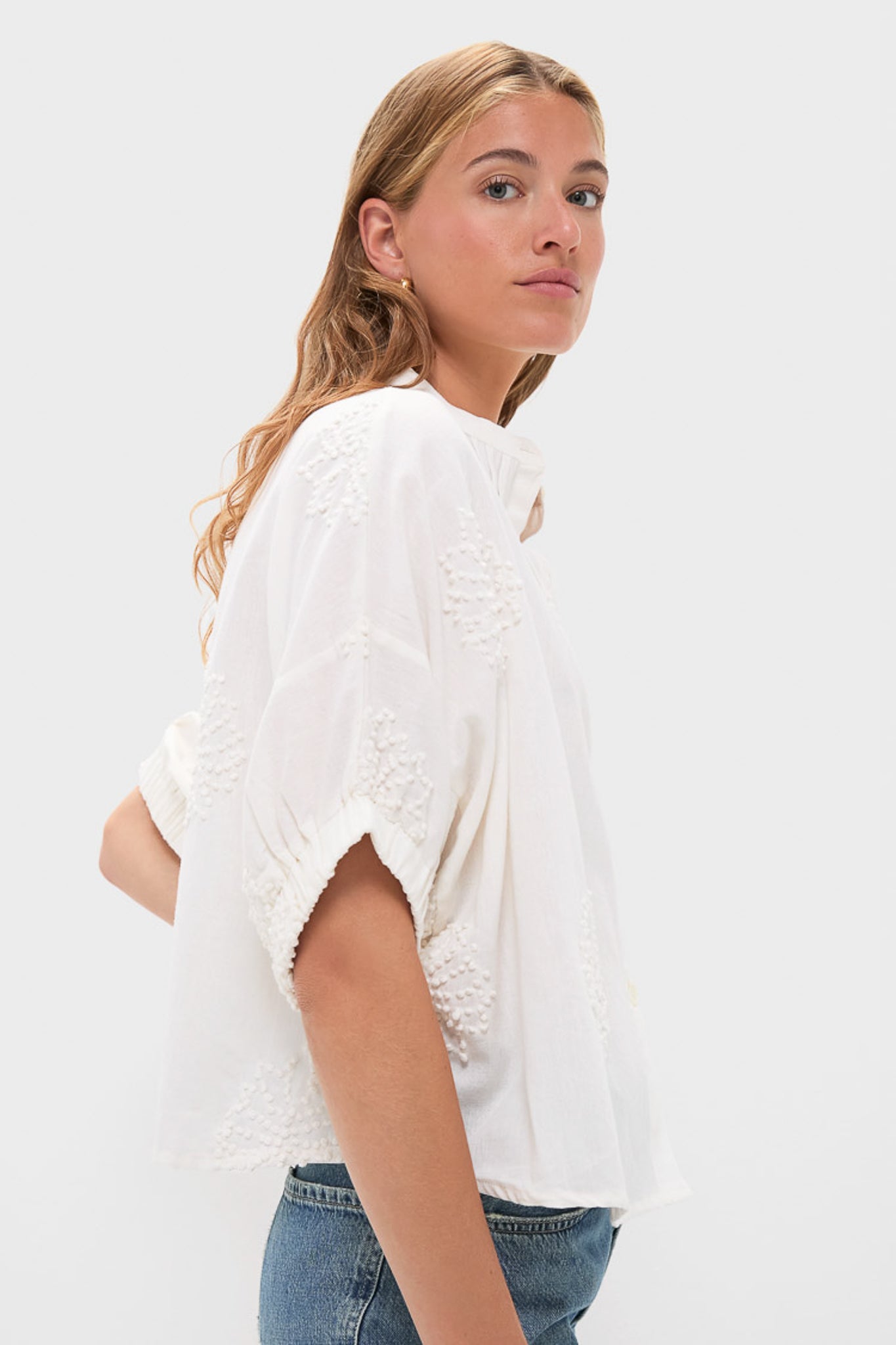 A woman with long, wavy blonde hair models a loose white embroidered cotton flax blouse from Love Tomas by Emerson Fry with blue jeans, posing sideways against a plain light background.