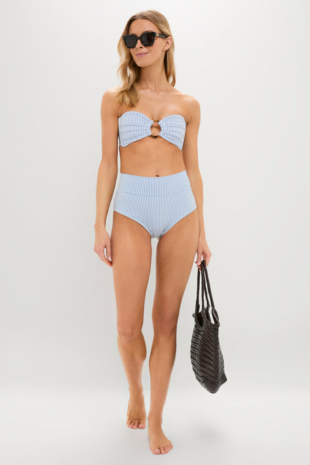 A woman models Montce’s blue and white striped bandeau bikini top with high-waisted bottoms, accessorized with a woven black bag and black sunglasses—effortlessly flattering swimwear.