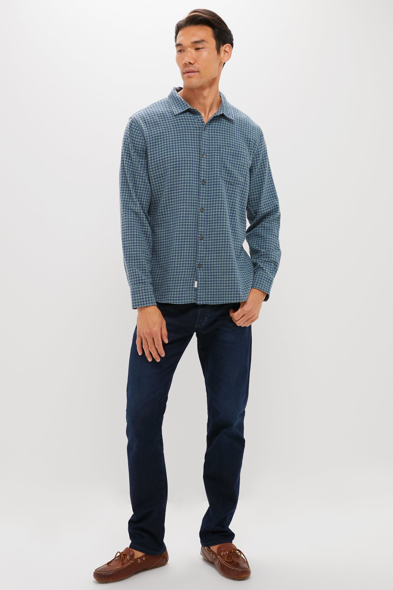 A man stands against a white background wearing Marine Layer’s lightweight gingham shirt, dark blue jeans, and brown boat shoes—ideal for those seeking sustainable closet staples.