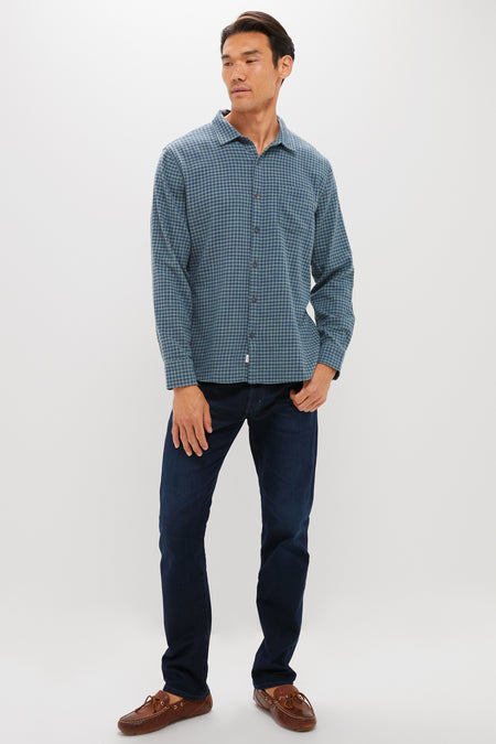A man stands against a white background wearing Marine Layer’s lightweight gingham shirt, dark blue jeans, and brown boat shoes—ideal for those seeking sustainable closet staples.