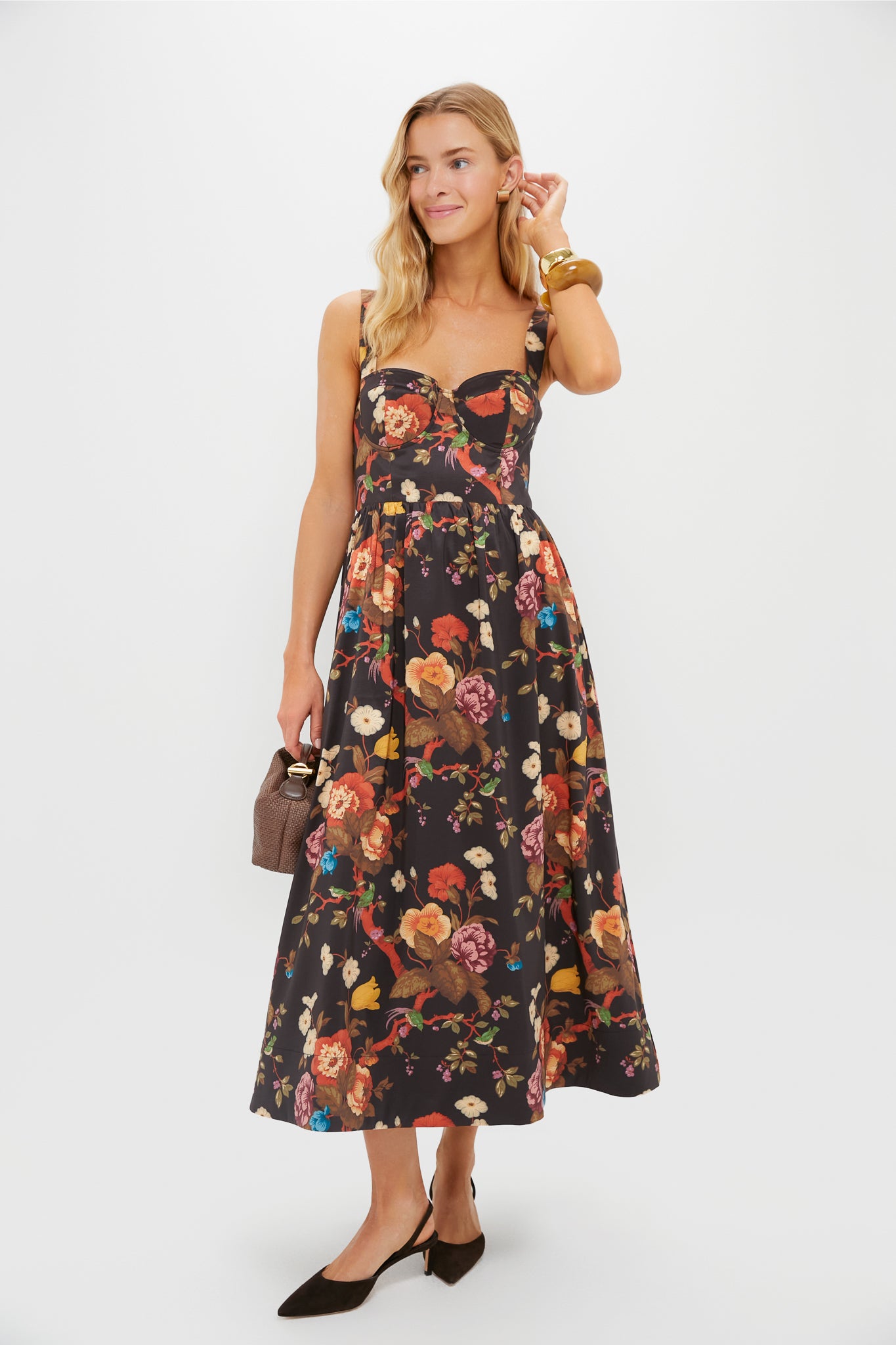 A woman wears a Tuckernuck Baroque Floral fit-and-flare midi dress with a sweetheart neckline, styled with a brown handbag, black heels, and gold bangles, posed against a plain white background.