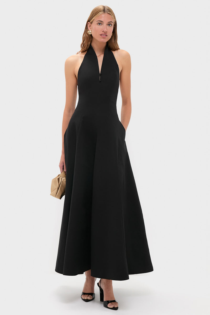 A woman in a STAUD sleeveless black maxi dress with a halter neckline, black heeled sandals, large earrings, and a beige clutch stands against a plain light background.