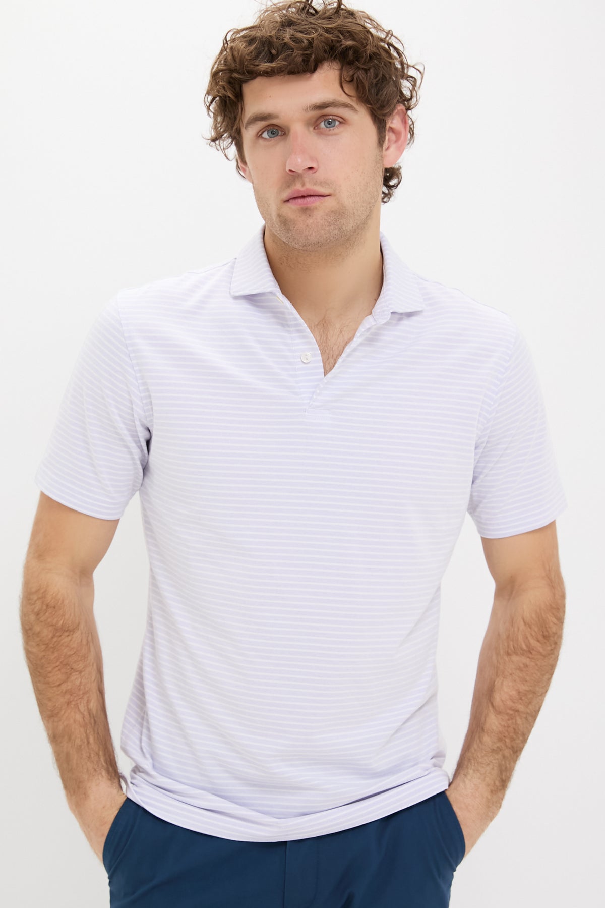A man with curly hair stands with his hands in his pockets, wearing a light purple Peter Millar polo and blue pants.