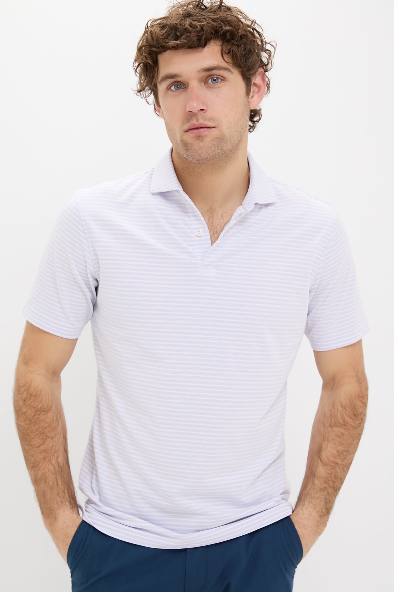 A man with curly hair stands with his hands in his pockets, wearing a light purple Peter Millar polo and blue pants.