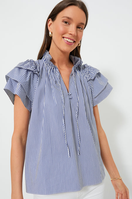 A woman wears the Hunter Bell Light Blue Stripe Millie Top—crafted from striped cotton shirting with flutter sleeves, a ruffled collar, and front ties—paired with white pants.