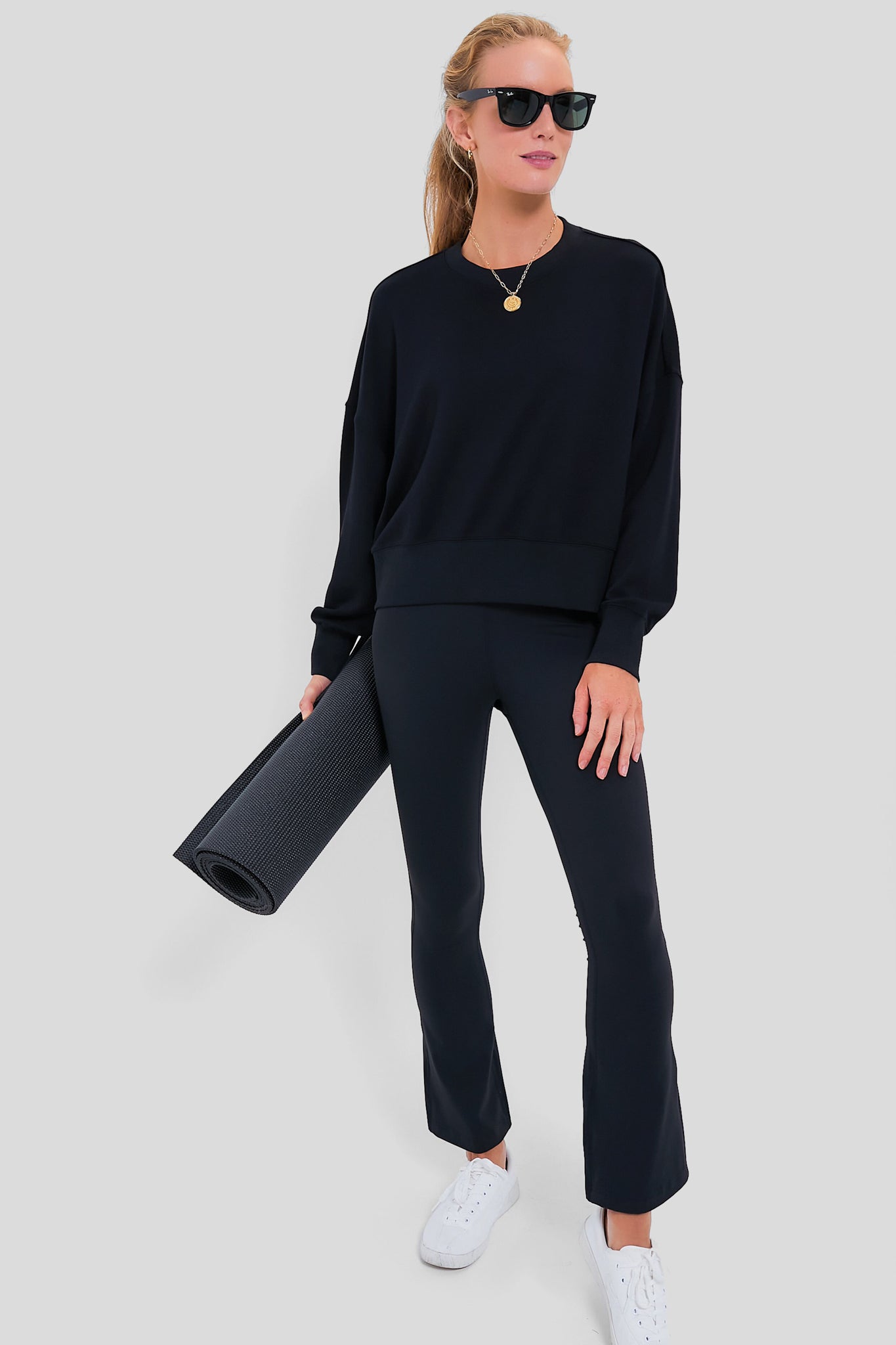 Woman models classic Black High Rise Compression Spandex Millie Yoga Pant; effortlessly paired with a loose sweatshirt, completing a fun, high-end athleisure look with rolled yoga mat.