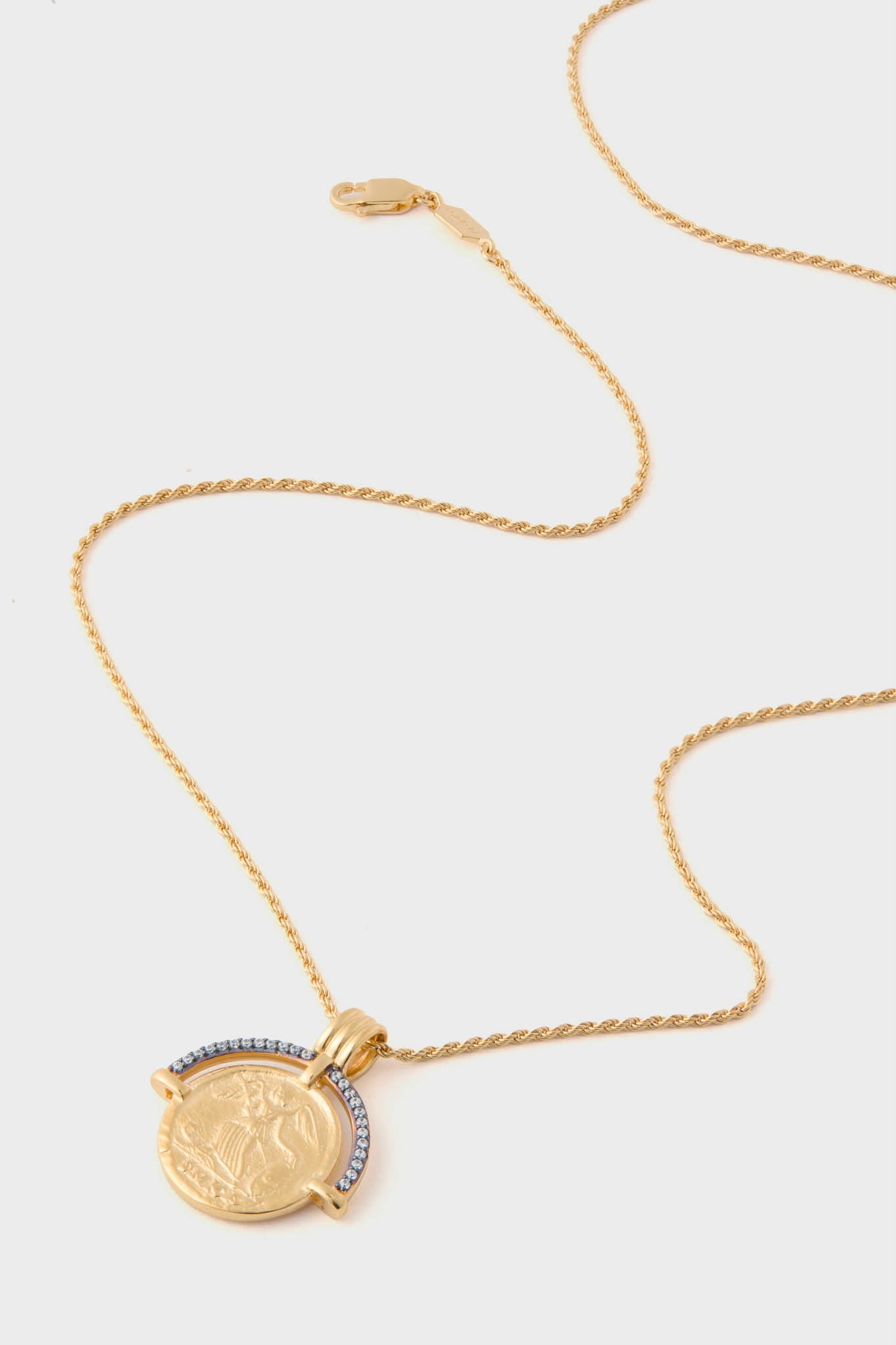 Missoma gold chain necklace with a round, coin-like pendant showcasing an embossed design and a row of clear stones along part of the edge.