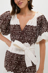 A woman wears THE GREAT. white vintage cotton dress featuring brown lace-like patterns, wide puff sleeves, a deep V-neckline, and a white waist belt tied in a bow.