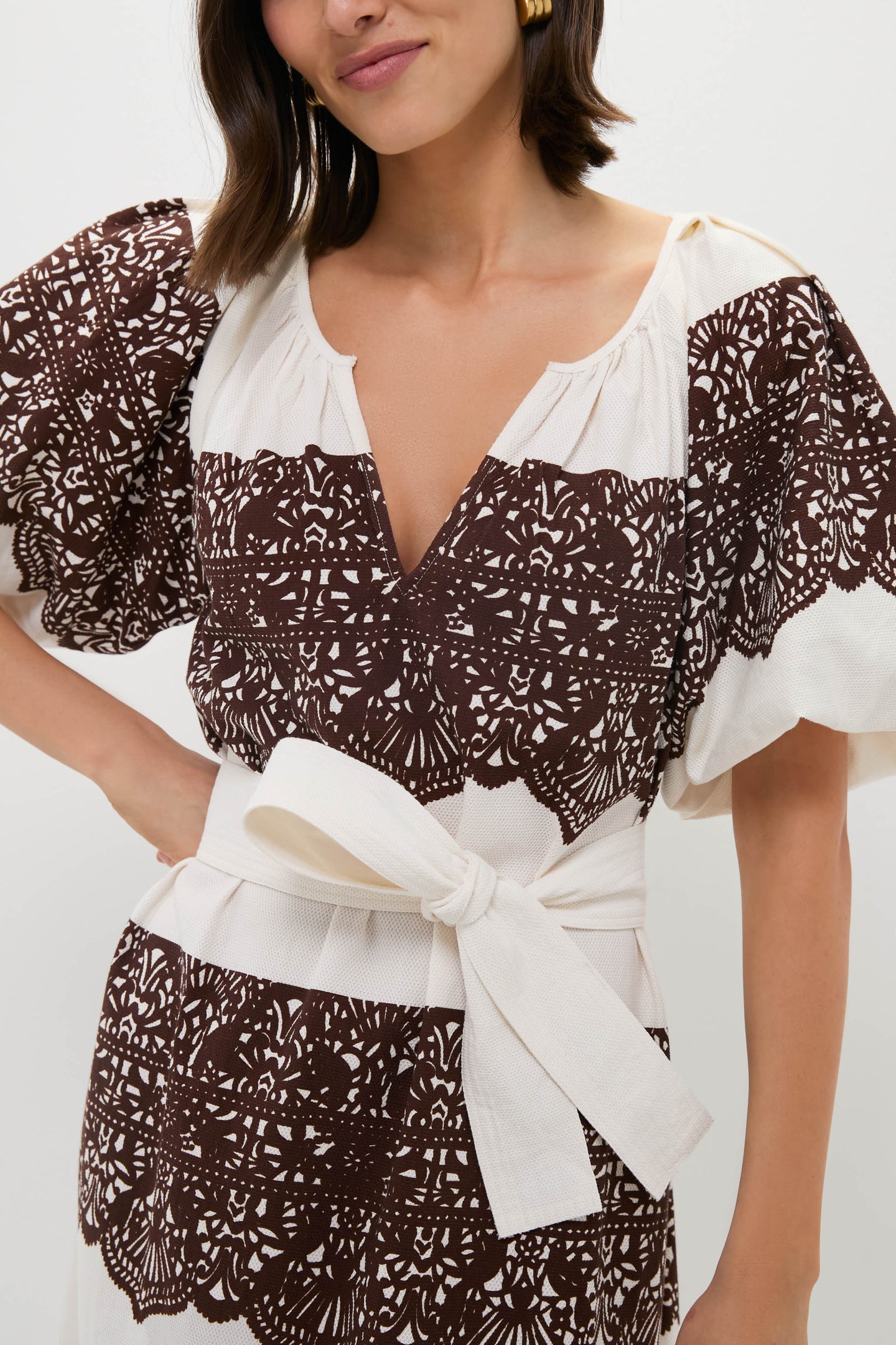 A woman wears THE GREAT. white vintage cotton dress featuring brown lace-like patterns, wide puff sleeves, a deep V-neckline, and a white waist belt tied in a bow.