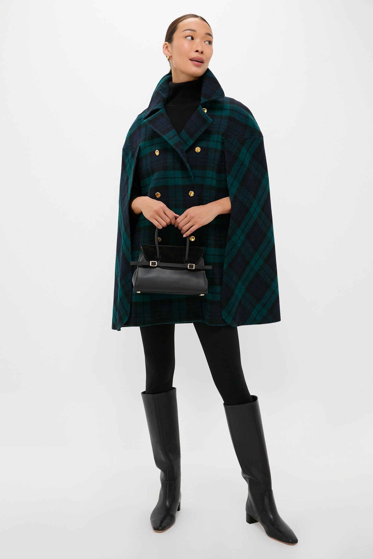 A person wearing Tuckernuck’s Blackwatch India Cape—a green and navy tartan coat—layered over a black turtleneck, black leggings, knee-high black boots, and carrying a small black handbag.