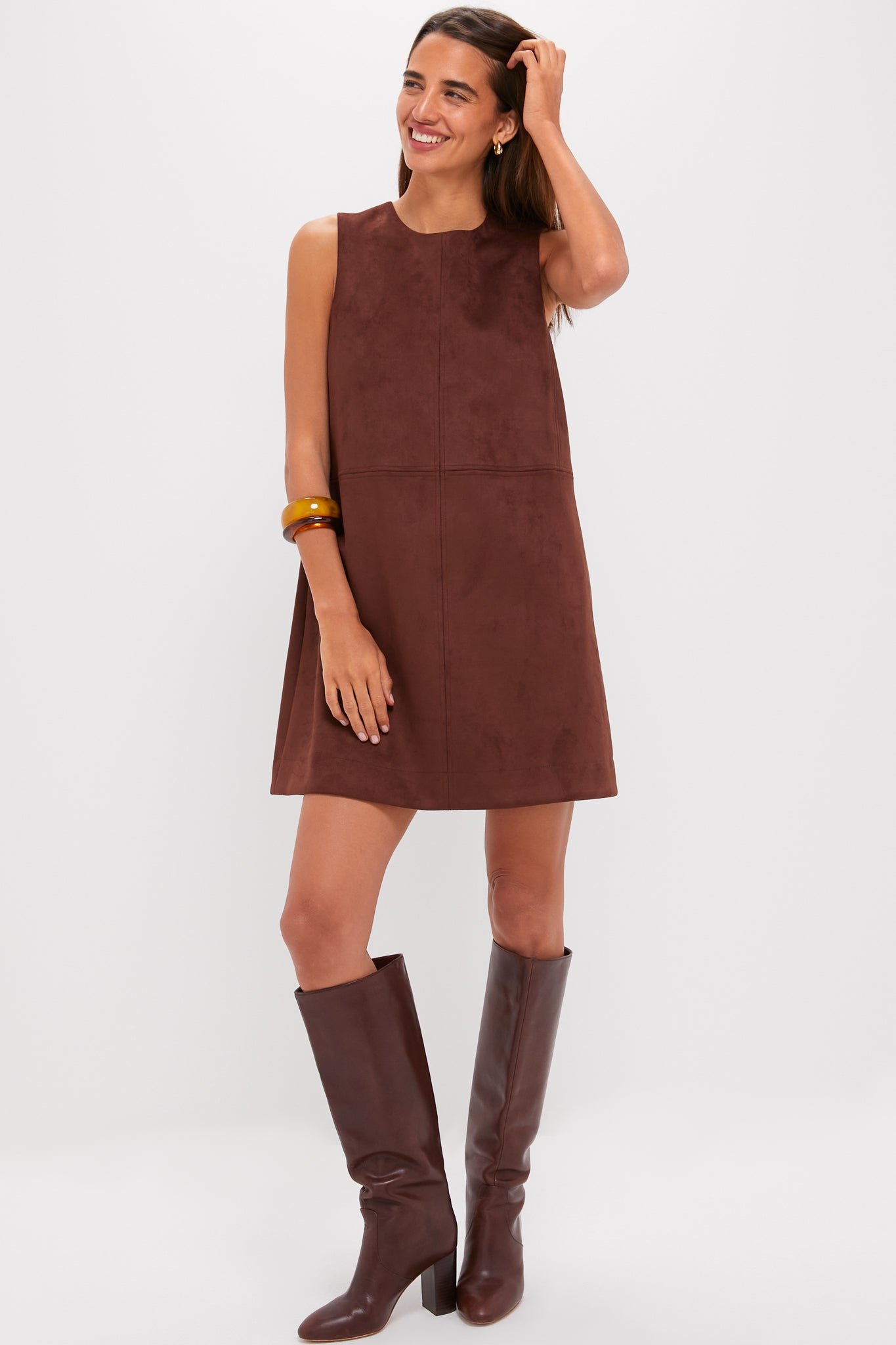 A woman wearing a Tuckernuck sleeveless vegan suede mini dress and knee-high brown boots stands against a white background, smiling with one hand in her hair.