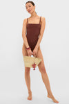 Woman in a classic Cappuccino Isabel One Piece swimsuit, holding a woven tote with coral charm. Effortless style with hoop earrings and chunky bangles. High-end, fun fashion.