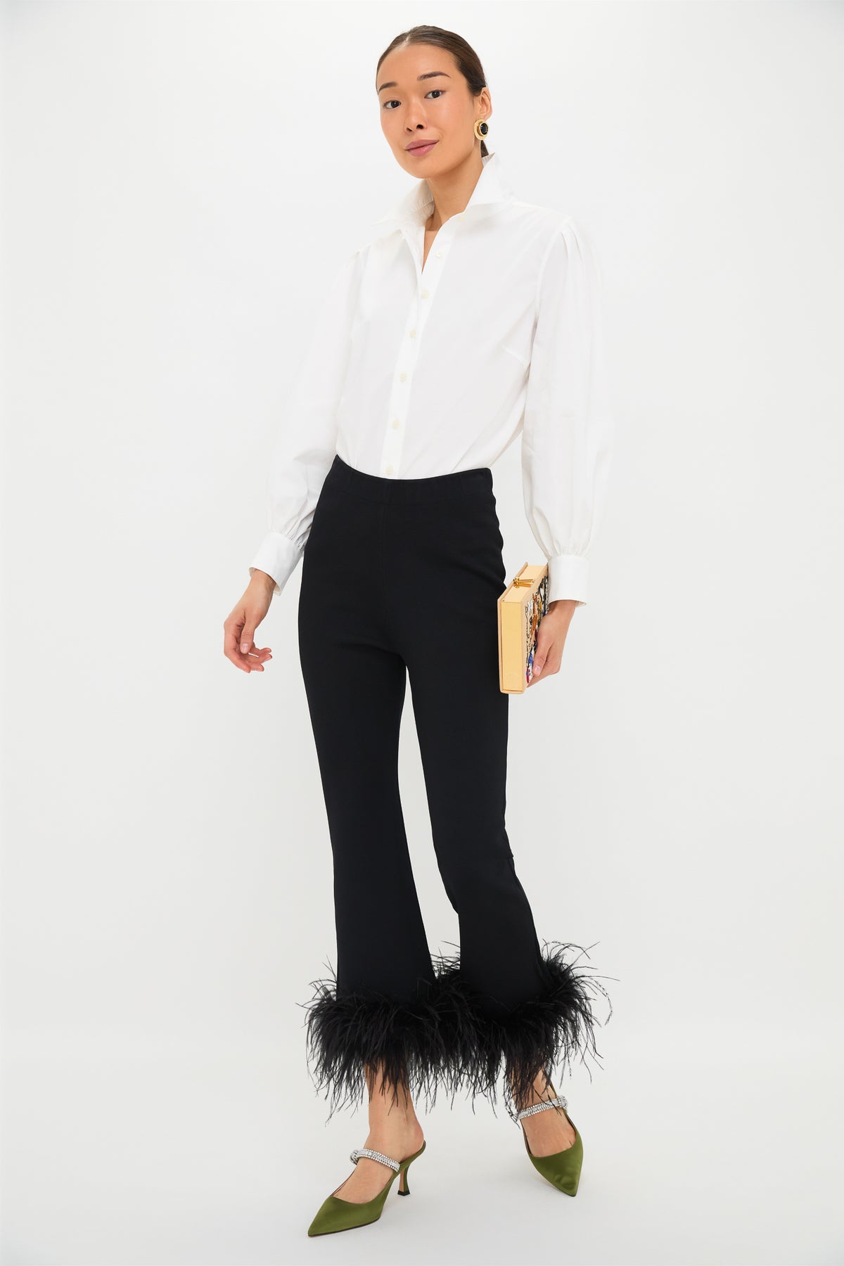 A person in a white button-up shirt, Tuckernuck Black Feather Knit Ashford Pants with feather-trimmed hem, green heels, and a beige clutch stands against a plain white background—these pants are perfect for making a statement at any party.