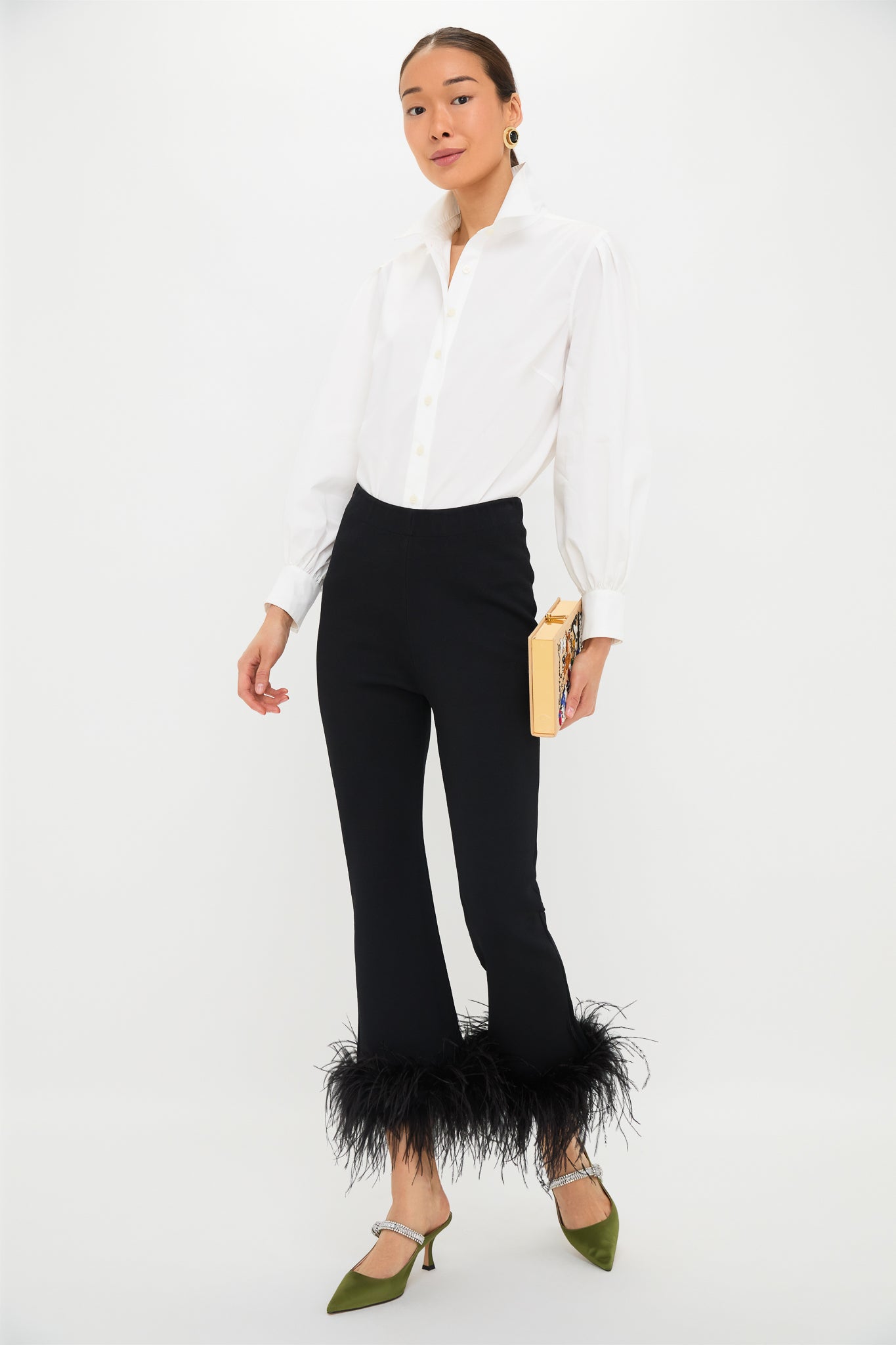 A person in a white button-up shirt, Tuckernuck Black Feather Knit Ashford Pants with feather-trimmed hem, green heels, and a beige clutch stands against a plain white background—these pants are perfect for making a statement at any party.