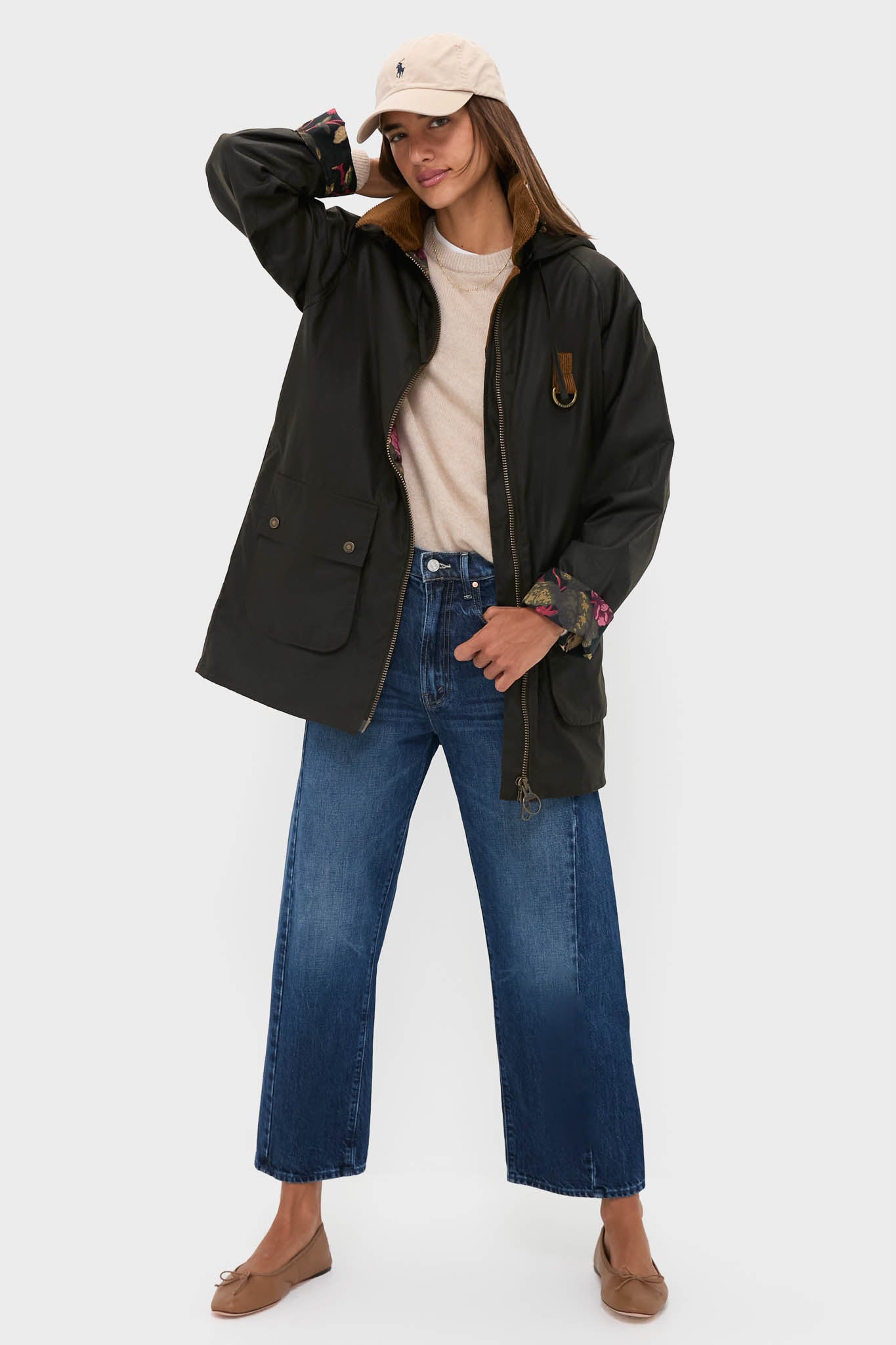 A woman wears a beige cap, light sweater, Barbour x Tuckernuck wax jacket, blue wide-leg jeans, and tan flats, standing with one hand on her head against a plain background.