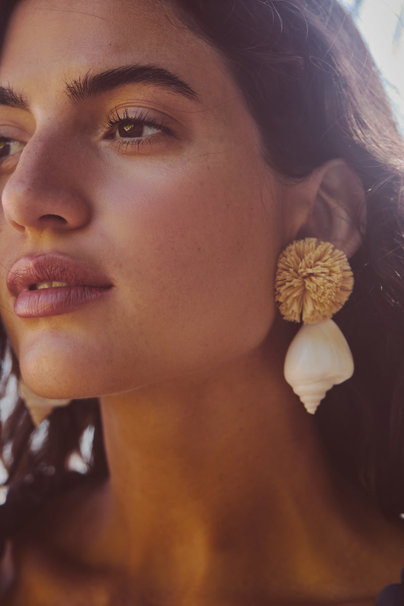 Young woman wearing Shell and Pom Elaine Earrings; featuring a fluffy raffia pom and spiral shell, exuding effortless, high-end coastal charm for a fun, classic look.