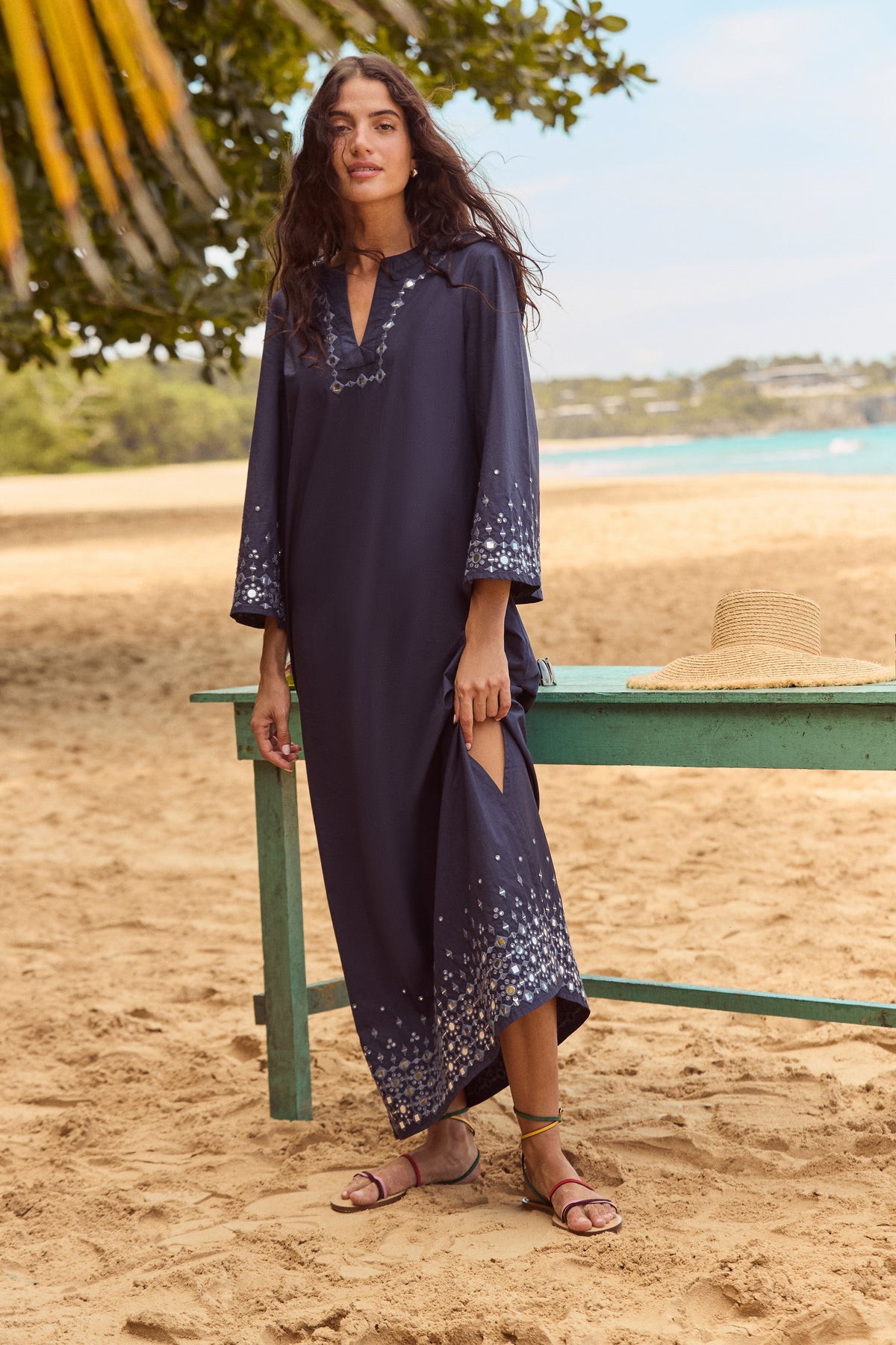 Woman in Navy Mirror Embellished Lorena Dress with embroidery, holding side slit on a beach beside a bench with straw hat, exuding effortless elegance and classic style.