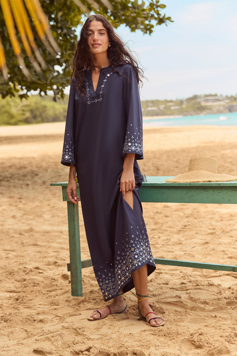 Woman in Navy Mirror Embellished Lorena Dress with embroidery, holding side slit on a beach beside a bench with straw hat, exuding effortless elegance and classic style.