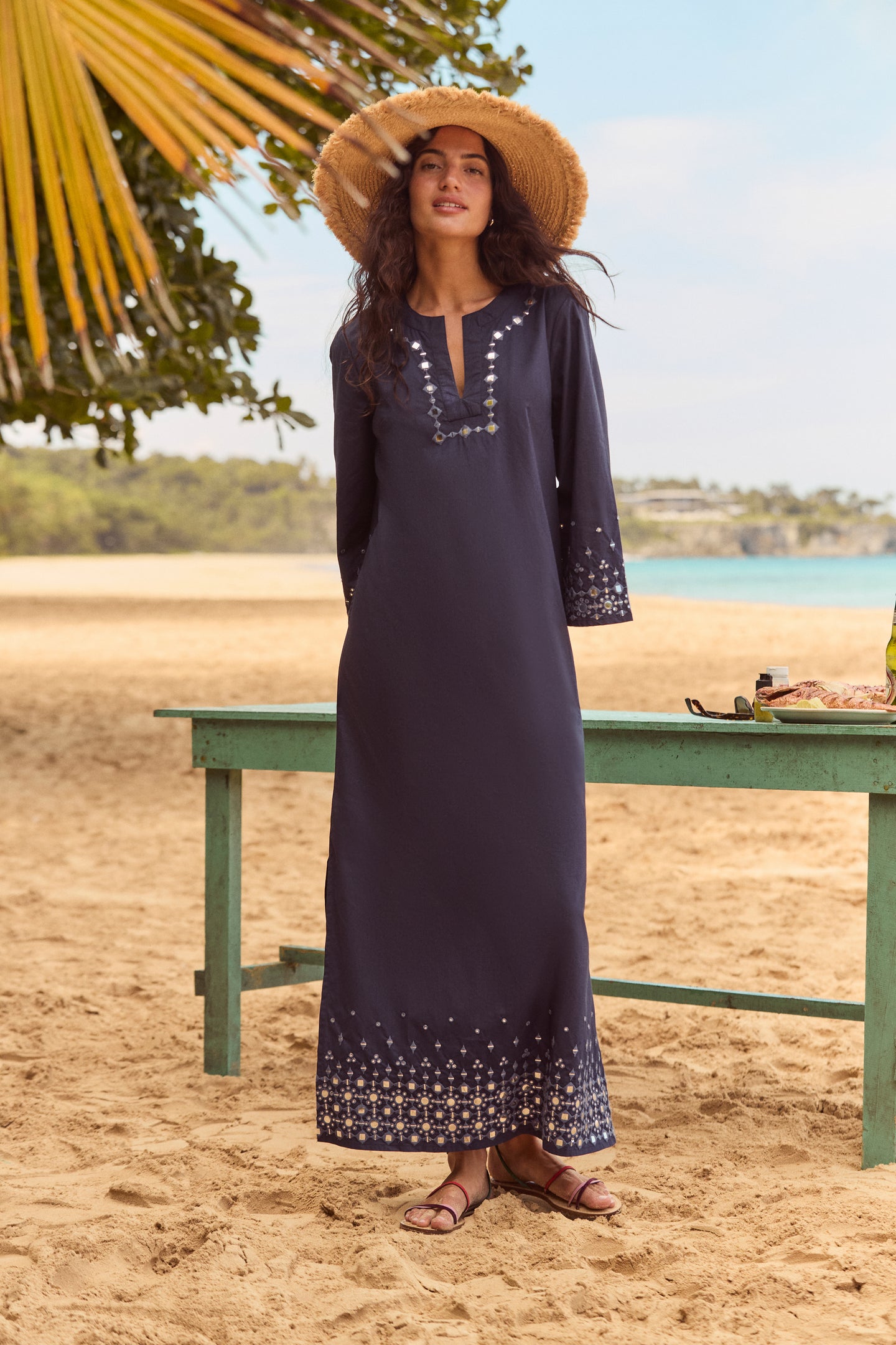 Woman in navy kaftan on beach wearing the Natural Raffia Fray Paloma Sunhat, showcasing its classic, effortless style with a frayed brim and secure neck ties.