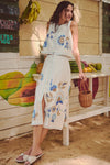 Woman in a classic, high-end Folk Flower Embroidered Sadie Skirt, embodying an effortless, fun style, holds a wicker basket at a beachside fruit stand.