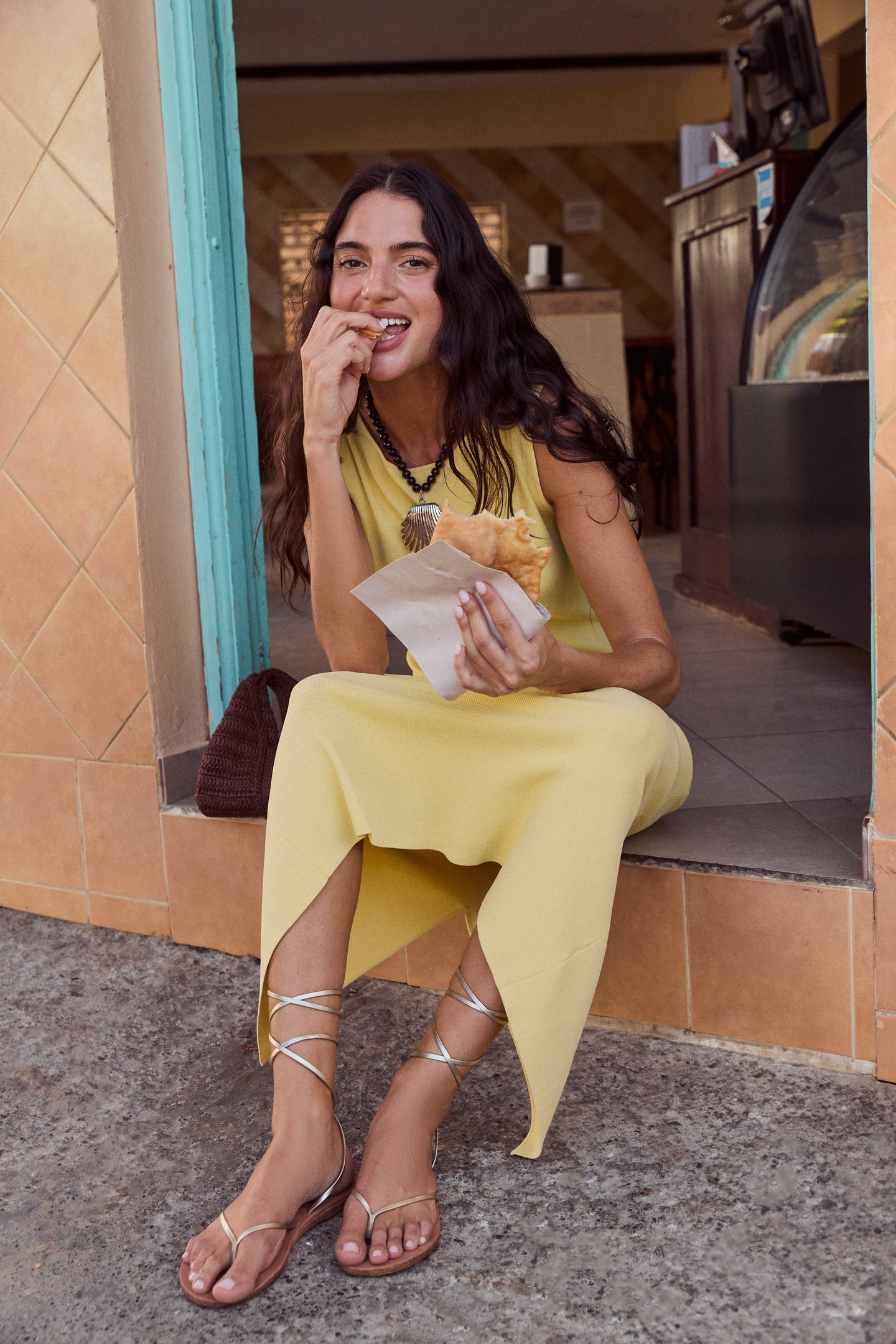 Woman enjoying a pastry at a café, effortlessly stylish in a yellow dress, accented by the classic, high-end Silver Holbox Necklace with bold beaded and shell details.