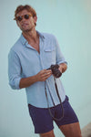 Effortlessly cool in a classic Light Blue Washed Denim Shirt, a man leans against a wall, embodying high-end casual style with a camera in hand.