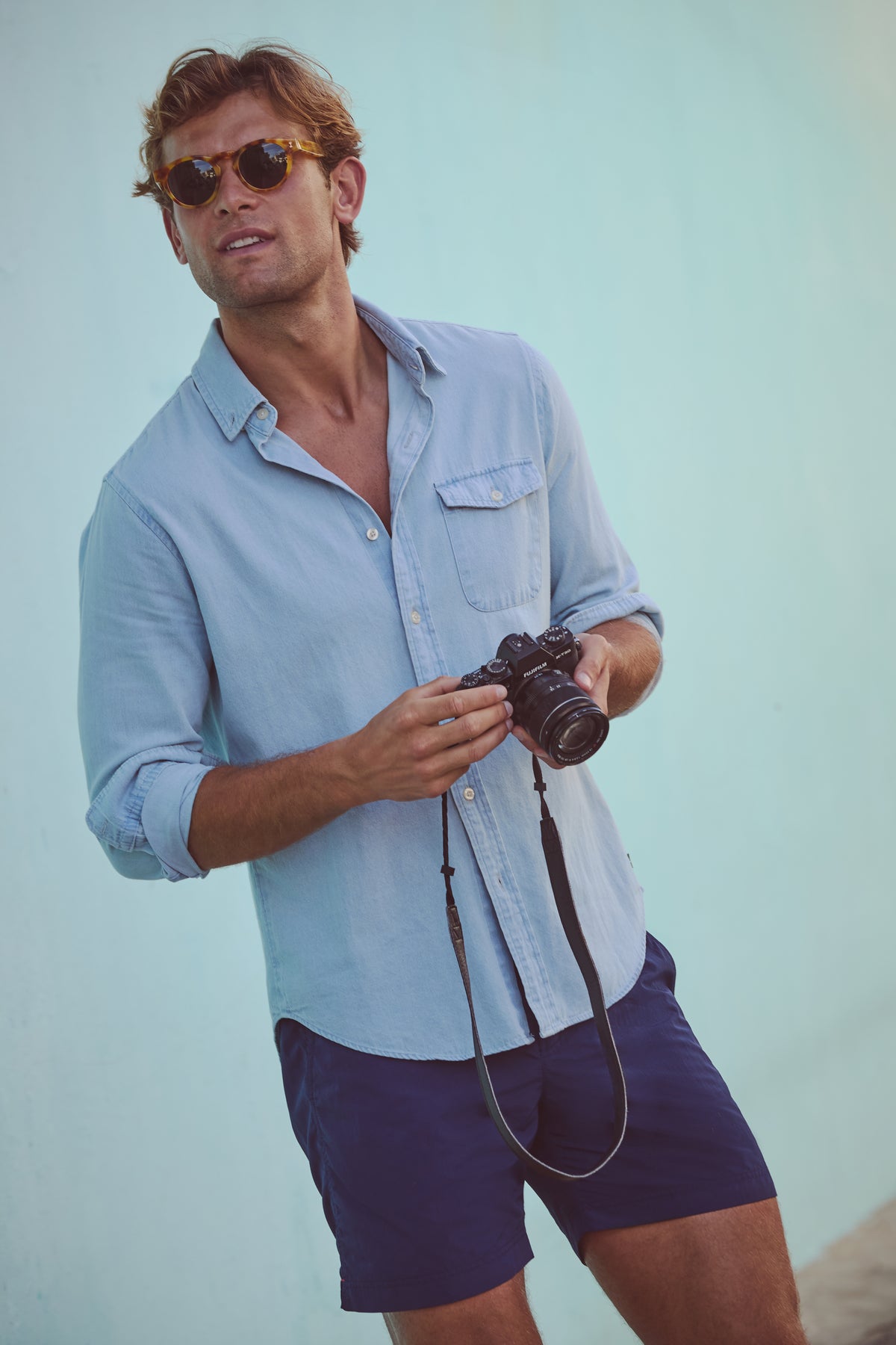Effortlessly cool in a classic Light Blue Washed Denim Shirt, a man leans against a wall, embodying high-end casual style with a camera in hand.
