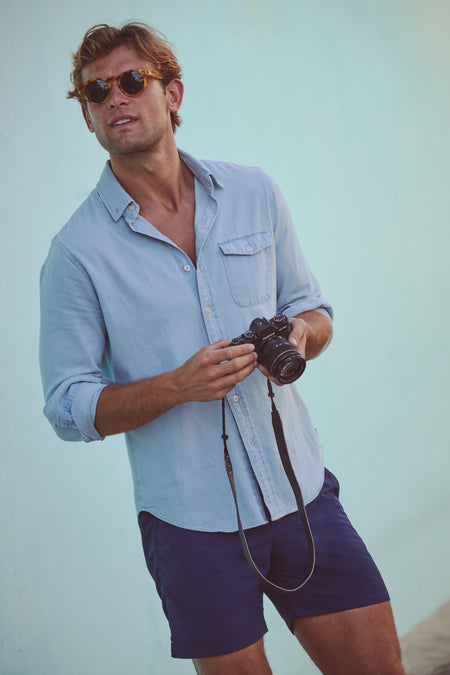 Effortlessly cool in a classic Light Blue Washed Denim Shirt, a man leans against a wall, embodying high-end casual style with a camera in hand.