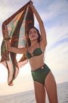 Young woman holds a patterned wrap above her head, showcasing the classic Sumatra Green Taita Bikini Bottom with high-rise, ruched detailing, and bead accent, embodying effortless, high-end beach style.