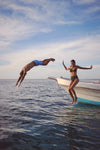 Two people leap off a boat, showcasing the classic, effortless style of Tropical Blue Bondi Snap Boardies, perfect for fun, high-end beach adventures.