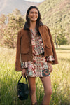 Woman in a two piece matching skirt set with a long sleeved top and mini skirt in colorful a floral pattern. Paired with a brown barn jacket, short cowboy boots, and a leather bag. 